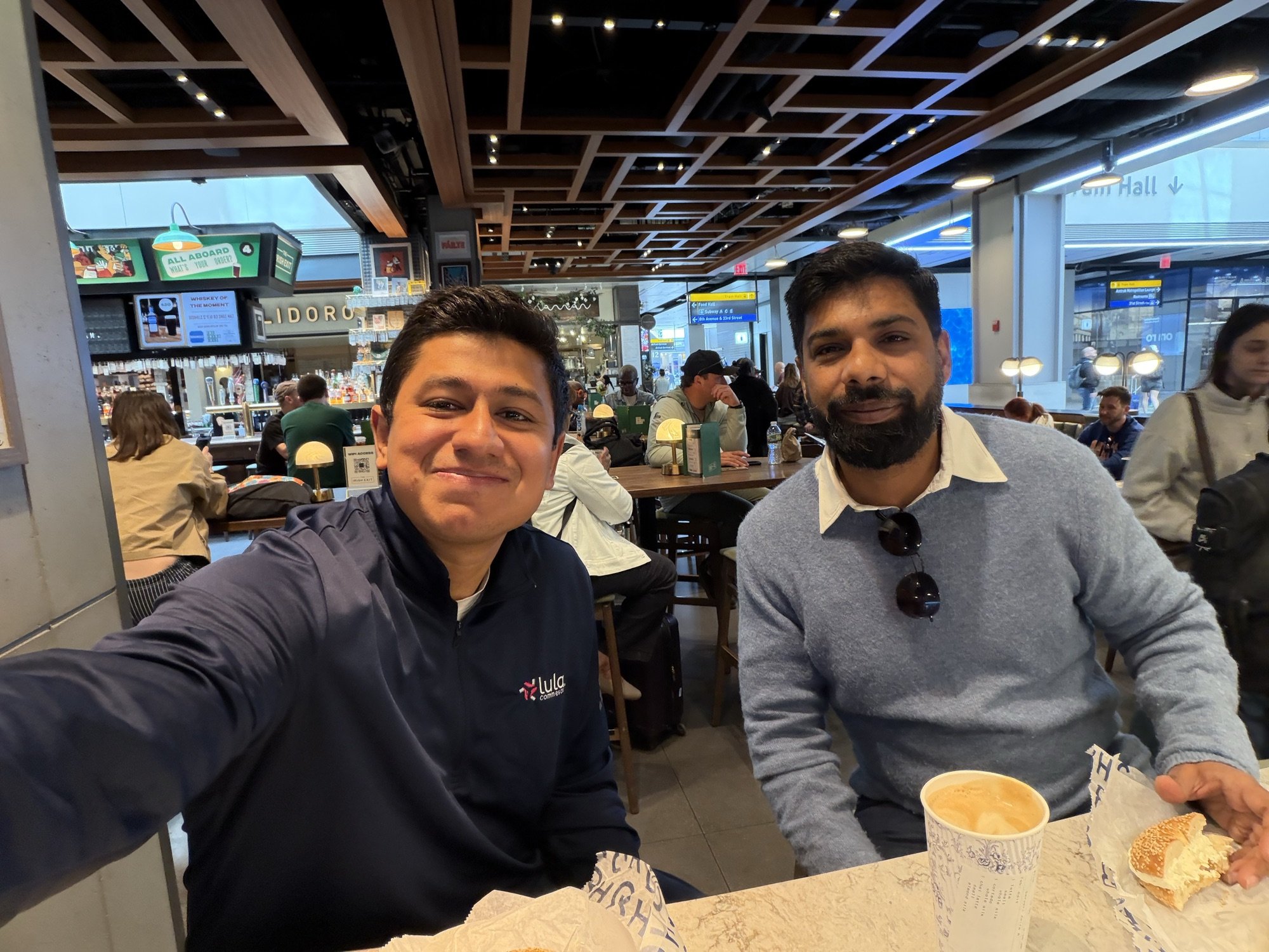 Two men seated together at a table in a busy food hall, taking a selfie.