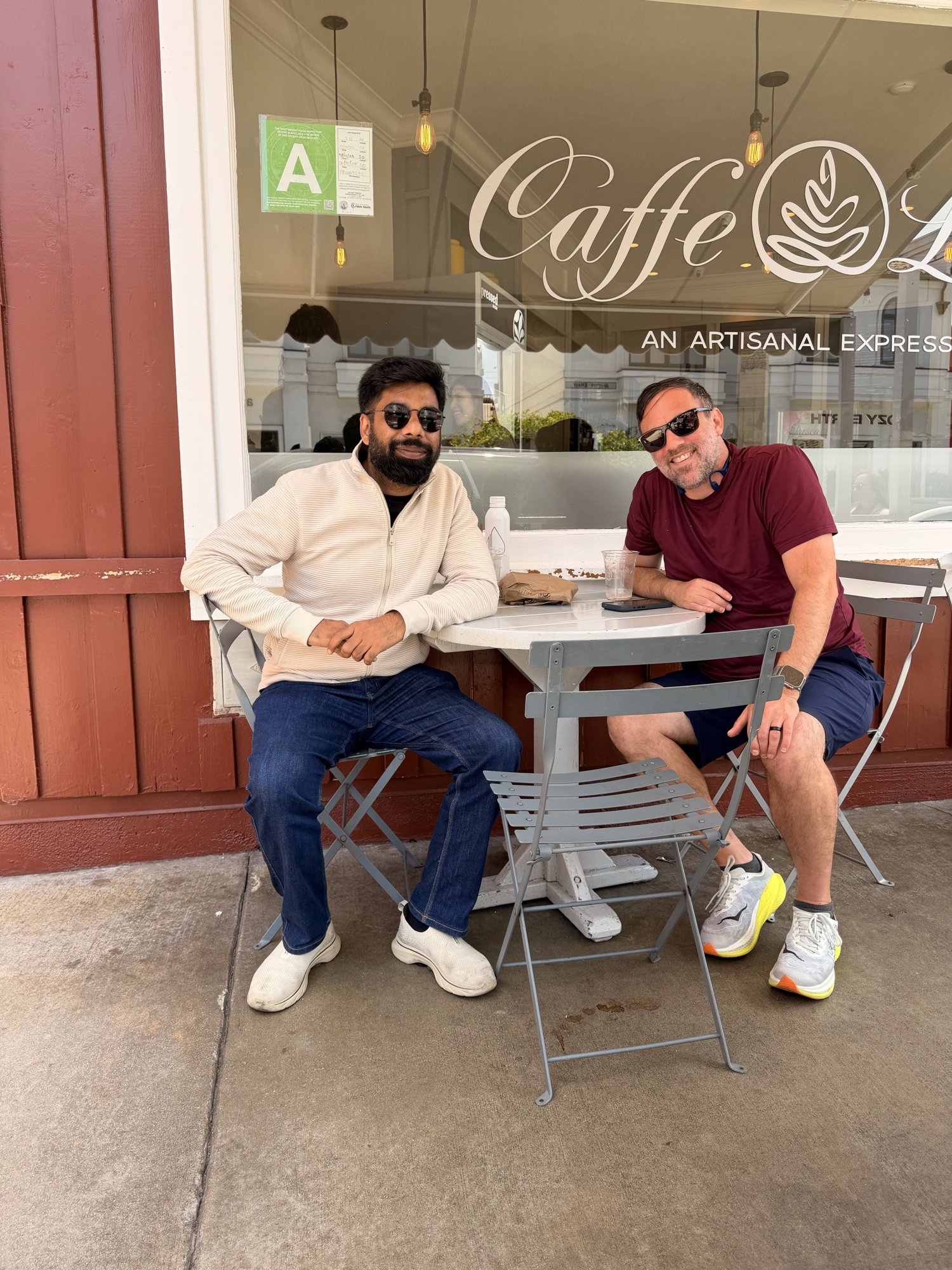 Two men wearing sunglasses seated together at an outdoor café table.