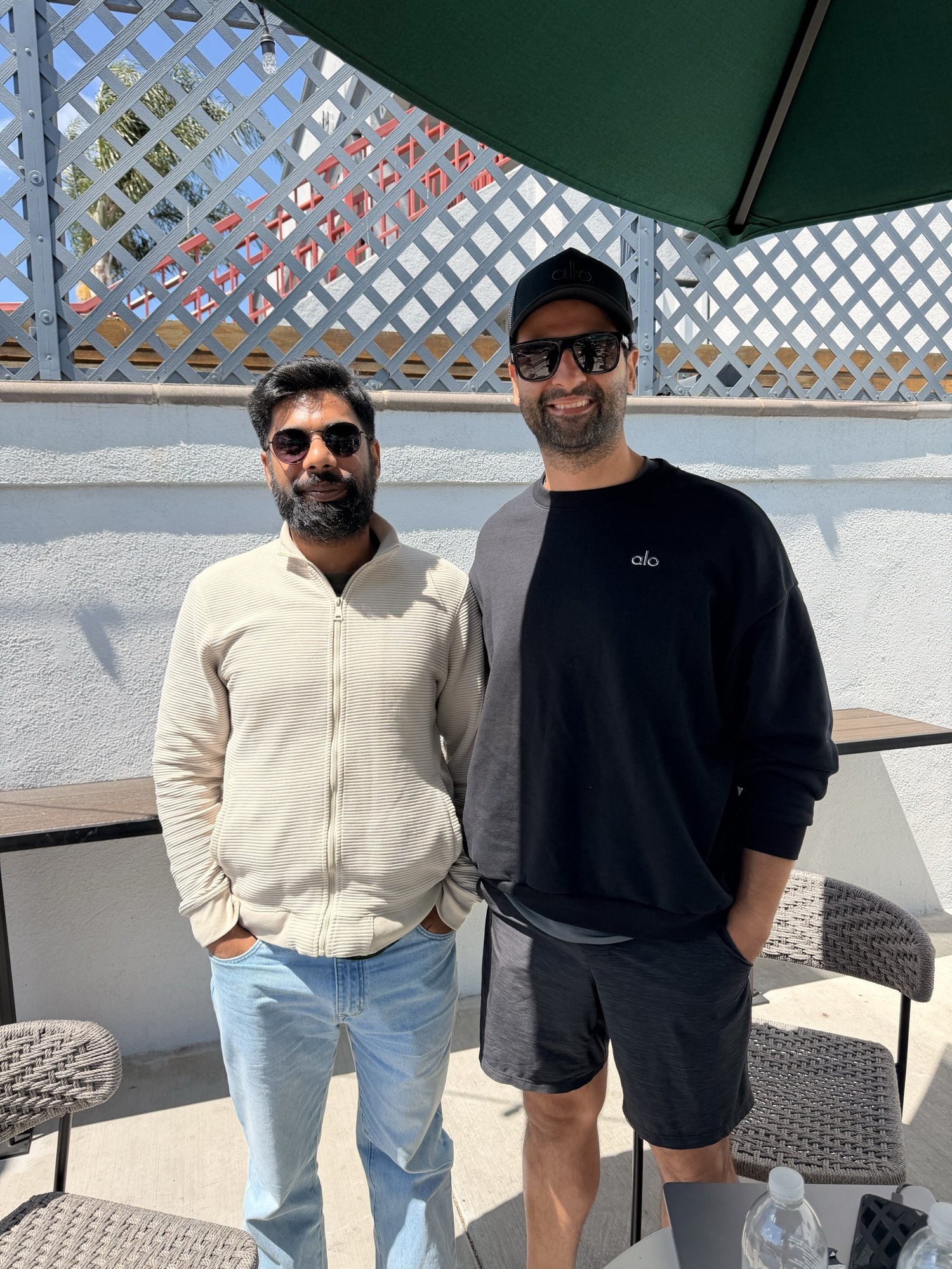 Two men wearing sunglasses standing together on an outdoor patio beneath a green umbrella.